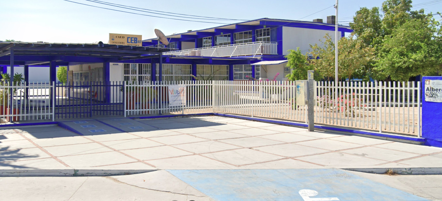 Fotografía en la que se muestra una explanada a la que se puede ingresar por medio de una rampa. Al fondo de la explanada hay una reja de color blanco que delimita la entrada y las instalaciones del plantel. Sobre una estructura techada hay un cartel que dice C E B 8, 2.